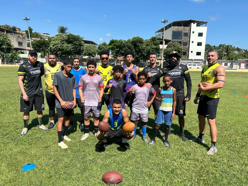 Atletas e treinadores do Tigres posam no campo com crianças e adolescentes do projeto social, segurando bolas de futebol americano